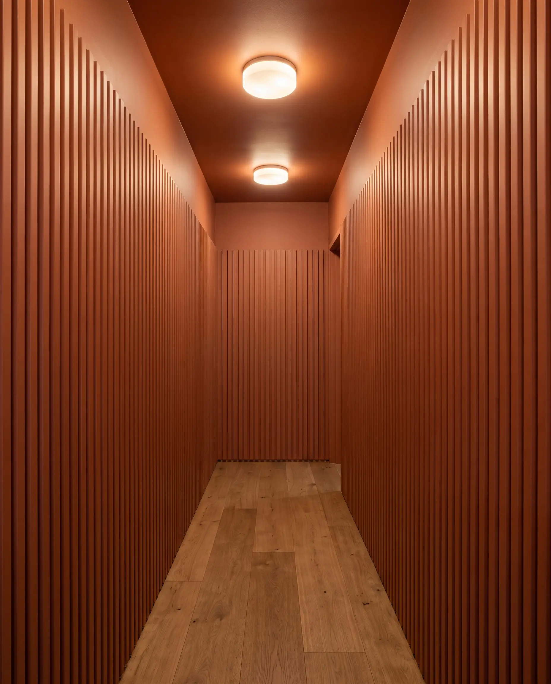 Deeply grooved vertical fluted 3D wall panels color-drenched in warm terracotta line a narrow hallway with minimalist oak flooring and flush-mount lighting.