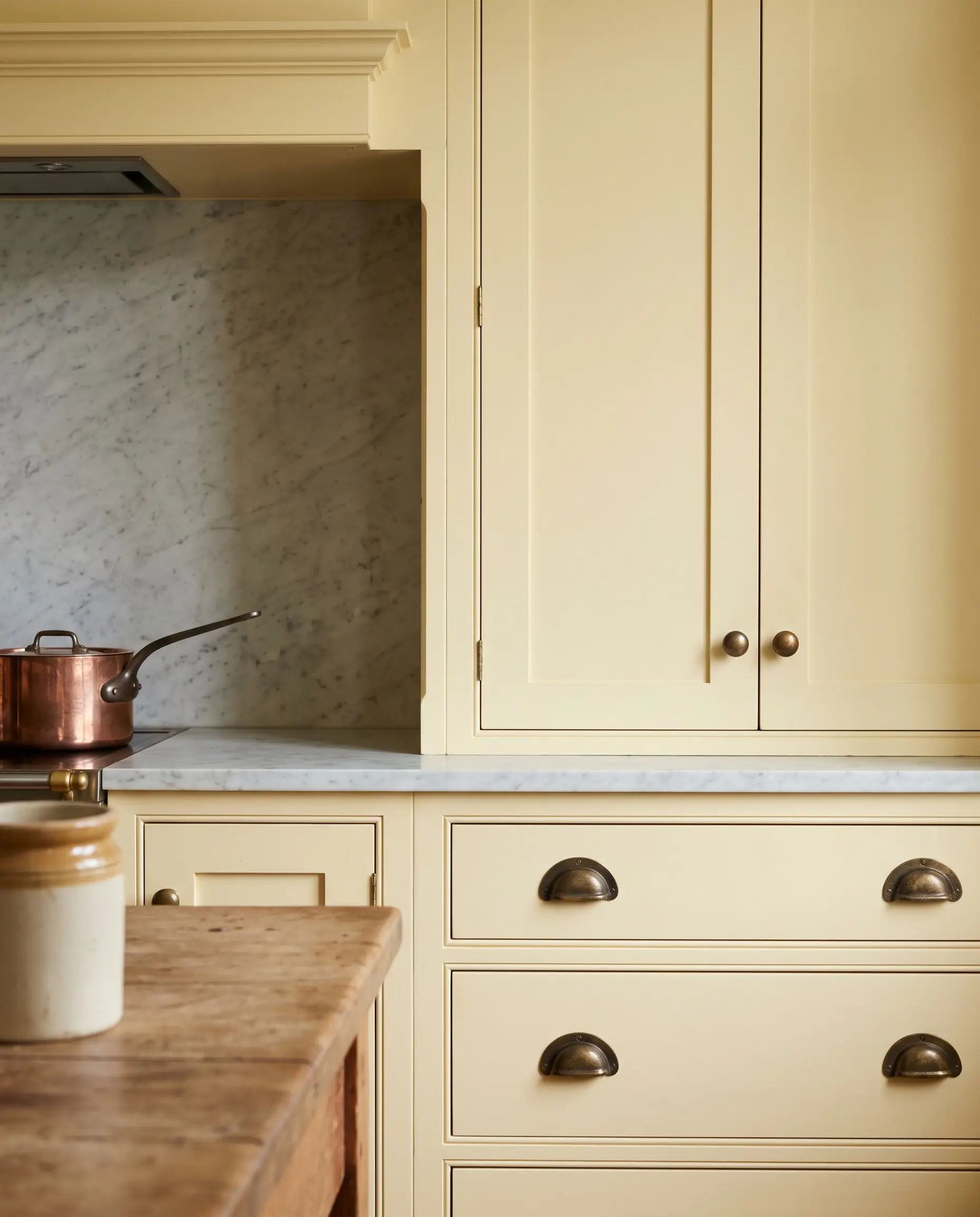Solid unlacquered brass cup pulls and ball knobs adorn butter-colored cabinetry set against a subtle marble backsplash in a well-appointed prep space.