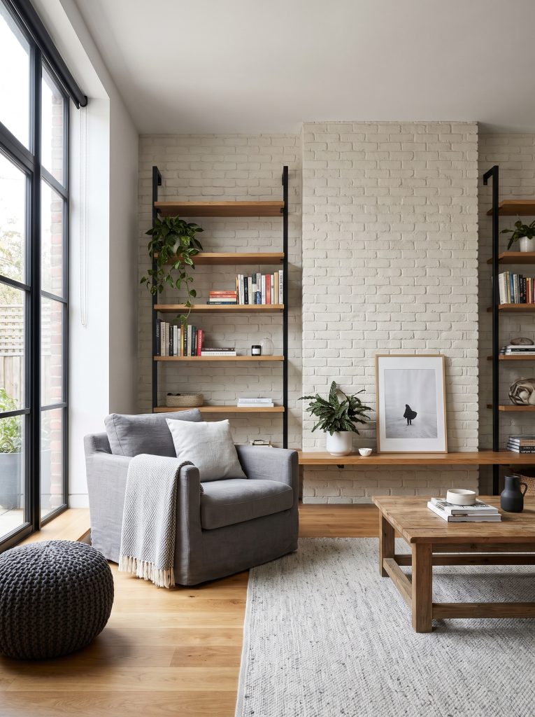Powder-coated matte black steel window frames and shelving brackets cut through a soft brick accent wall painted White Dove above warm oak flooring and a gray armchair.