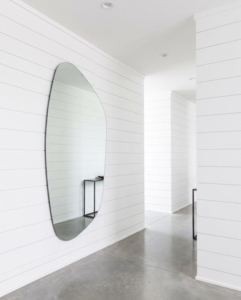 Horizontal deep-channel nickel gap shiplap painted bright monolithic white contrasts against an oversized organically shaped asymmetrical mirror mounted in a crisp modern hallway.