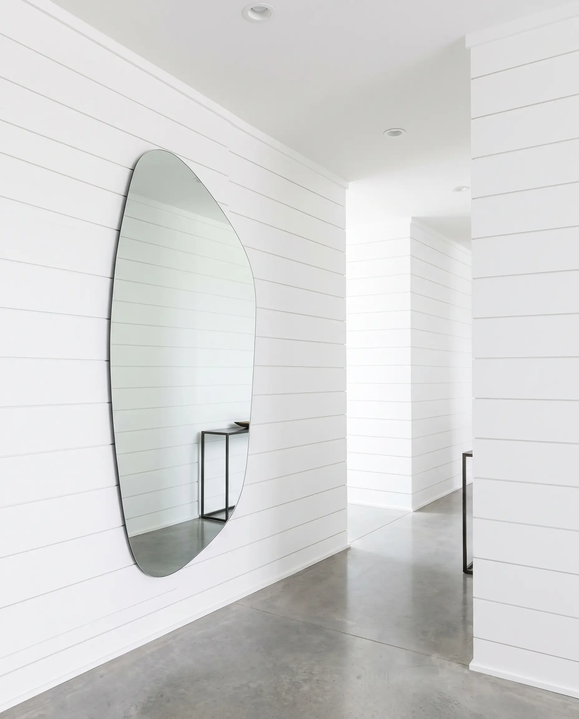 Horizontal deep-channel nickel gap shiplap painted bright monolithic white contrasts against an oversized organically shaped asymmetrical mirror mounted in a crisp modern hallway.