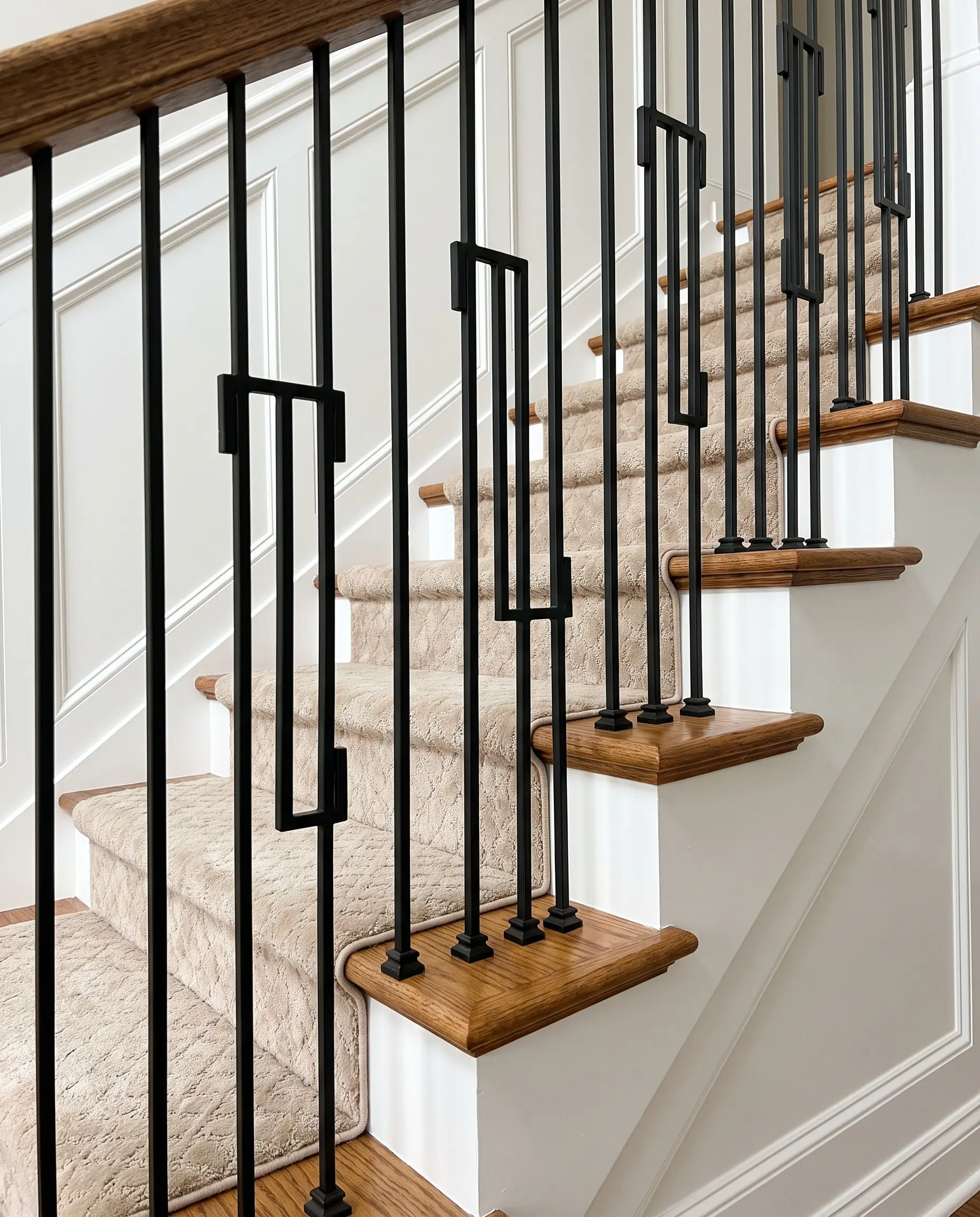 Hollow square matte black iron balusters with flat shoes line a crisp white painted stringer and warm oak treads covered by a neutral stair runner.