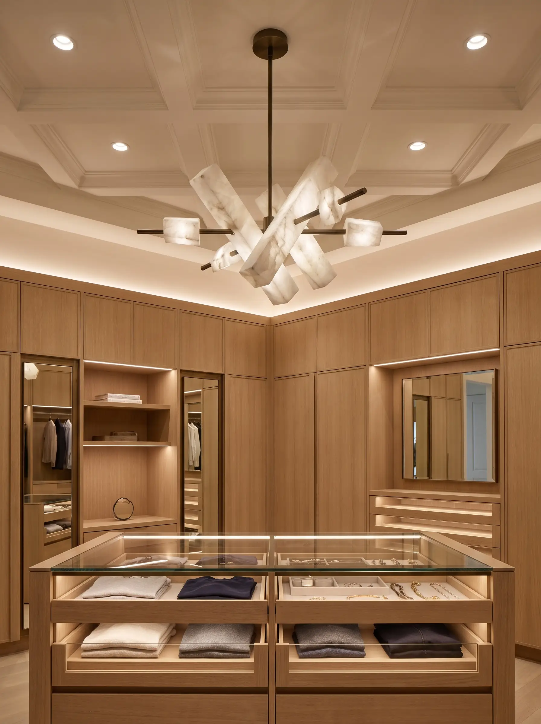 A sculptural natural alabaster stone and unlacquered brass chandelier hangs above a tempered glass center island reflecting 2027 closet and wardrobe design trends.