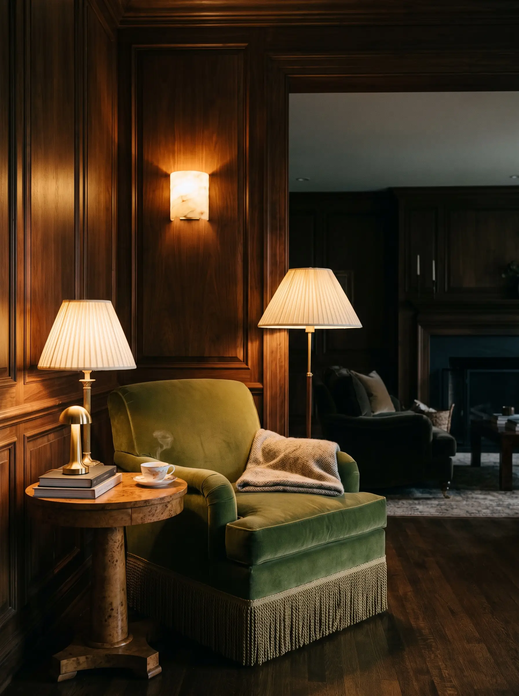 A glowing alabaster wall sconce and a pleated silk table lamp on a burl wood side table illuminate a heavy velvet fringed armchair and dark walnut paneled walls.