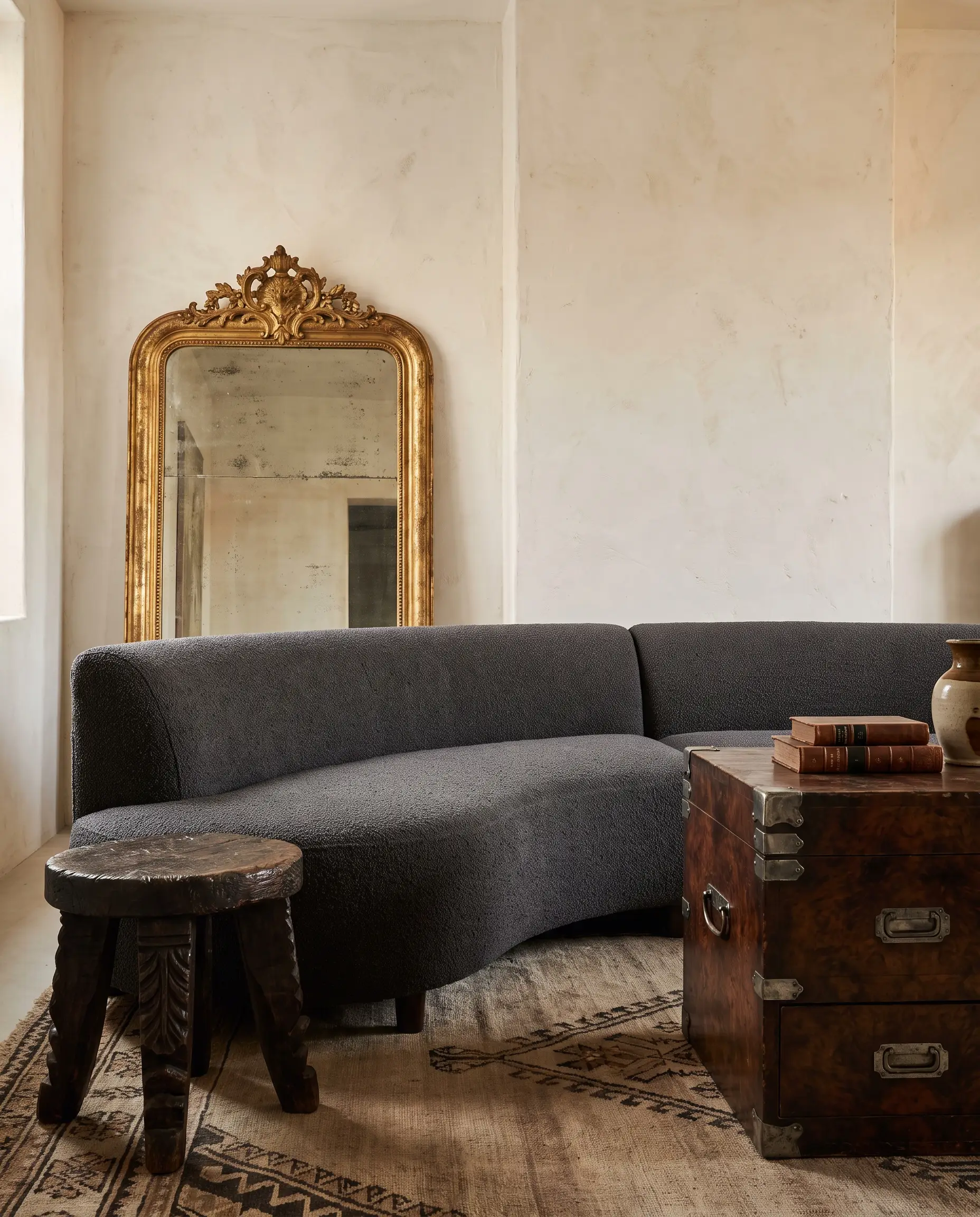 An oversized antique gilt mirror leans against a hand-troweled plaster wall beside a low-profile modern crescent sofa and a heavily patinated burl walnut campaign chest.