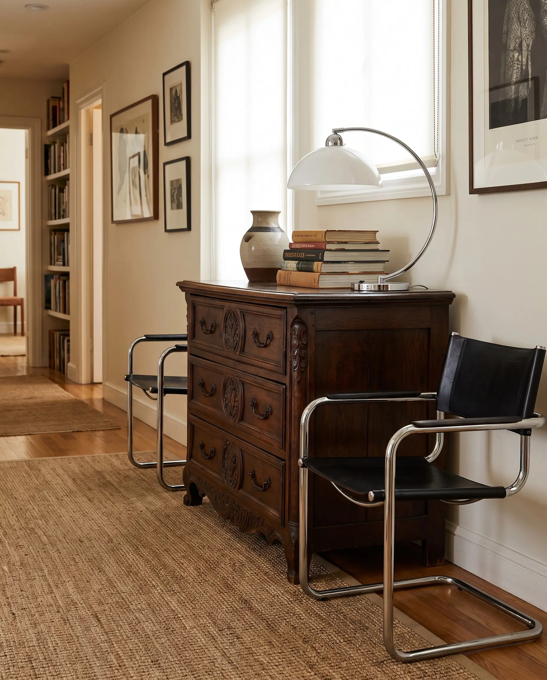 An ornately carved antique mahogany chest of drawers sits between tubular chrome 1970s Marcel Breuer chairs in a curated heritage hallway.