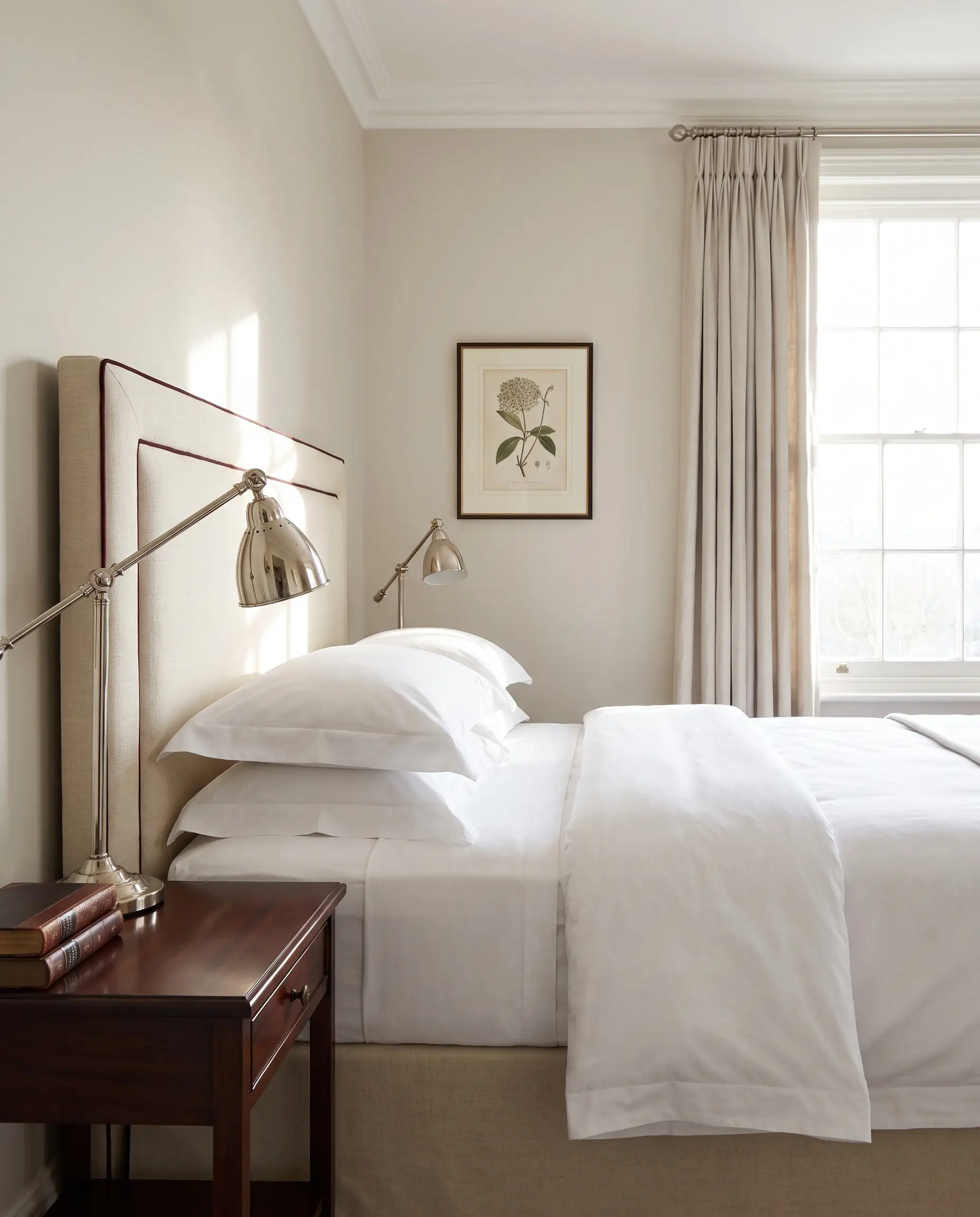 A crisp cream Belgian flax linen headboard outlined in dark oxblood contrast piping pairs with a polished nickel reading lamp and mahogany side table.