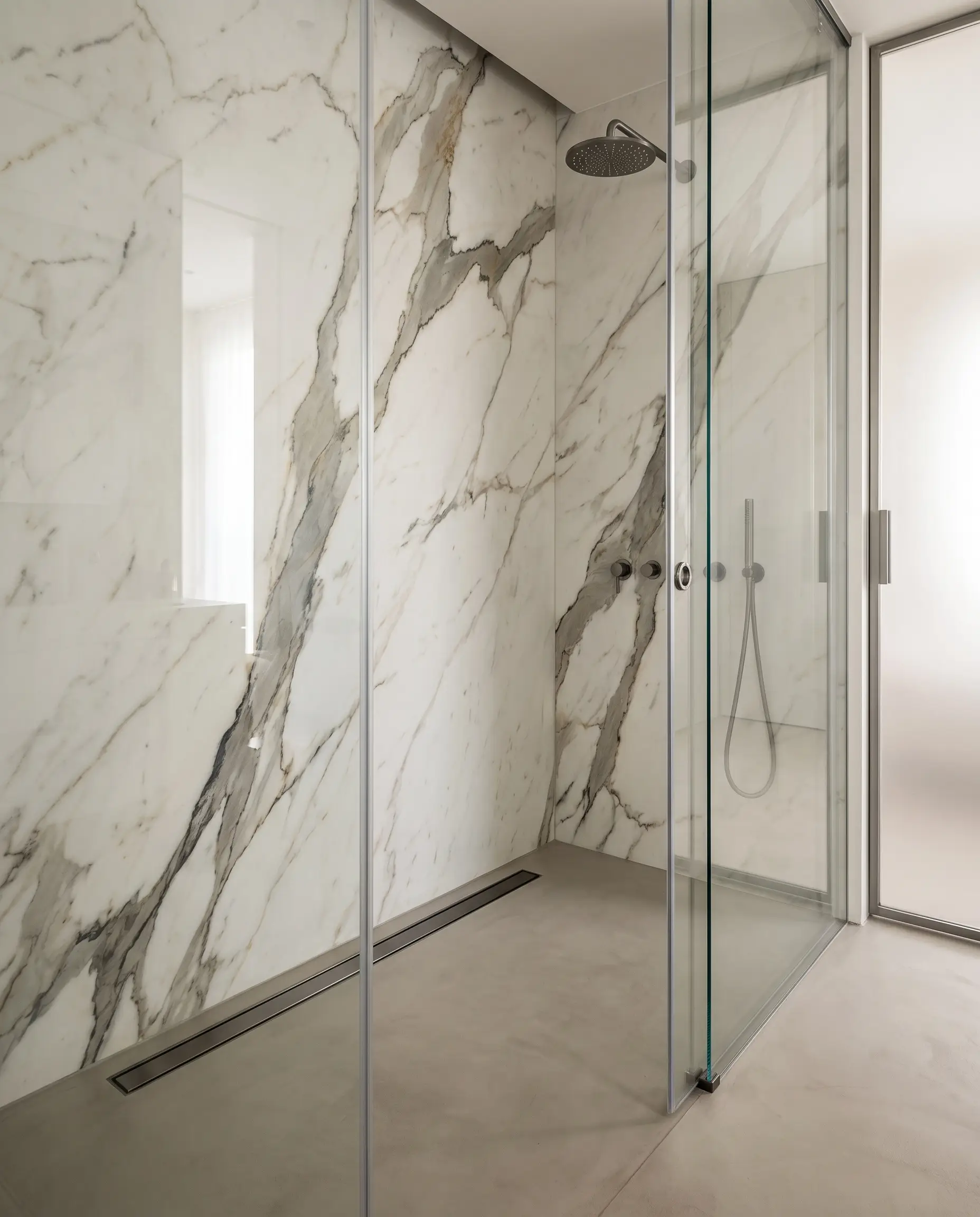 Massive floor-to-ceiling sintered stone slabs with dramatic Calacatta marble veining line a luxury shower enclosure featuring a sleek gunmetal linear drain.