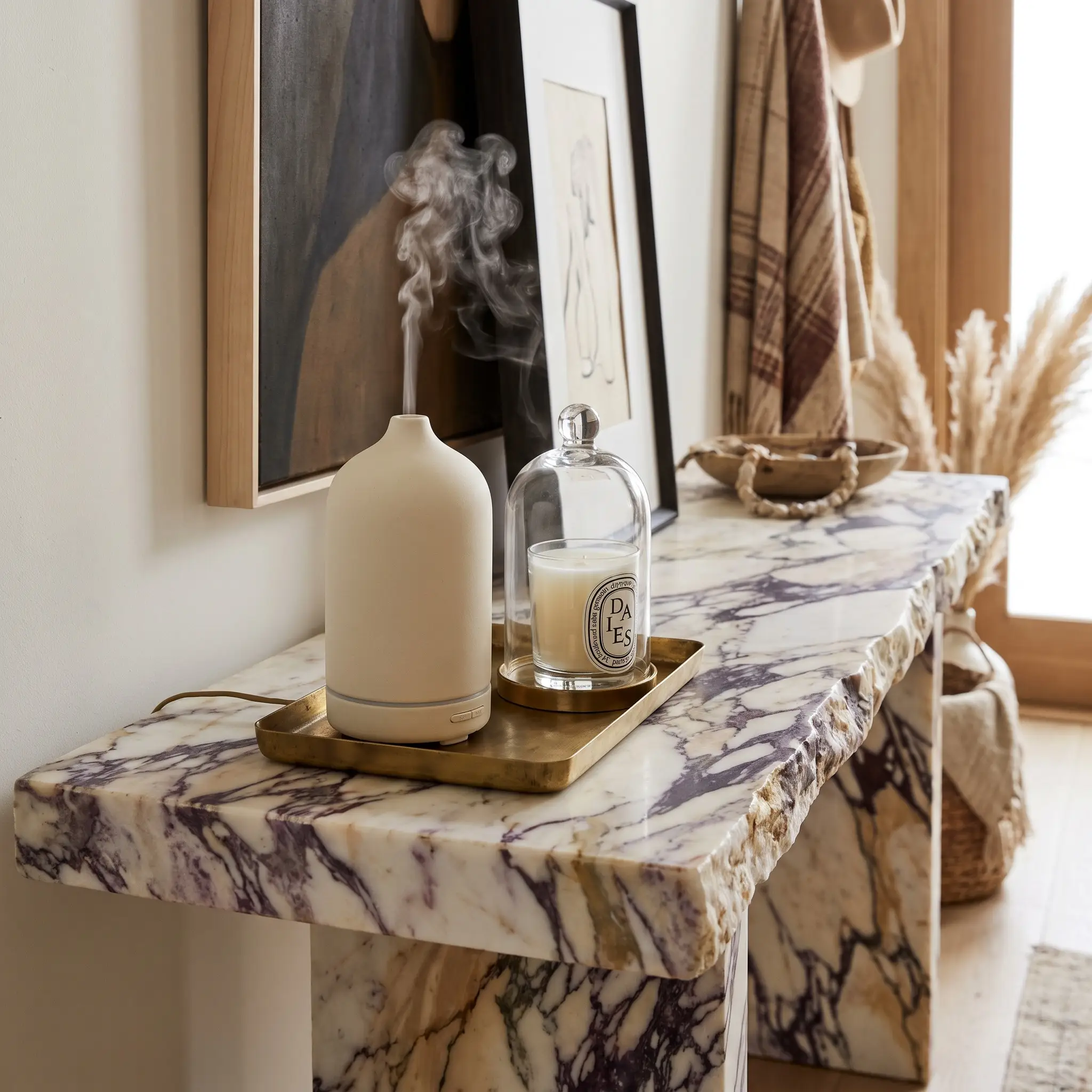 A matte ceramic diffuser and glass-cloched candle sit on a small unlacquered brass tray atop a heavily veined Calacatta Viola marble console.
