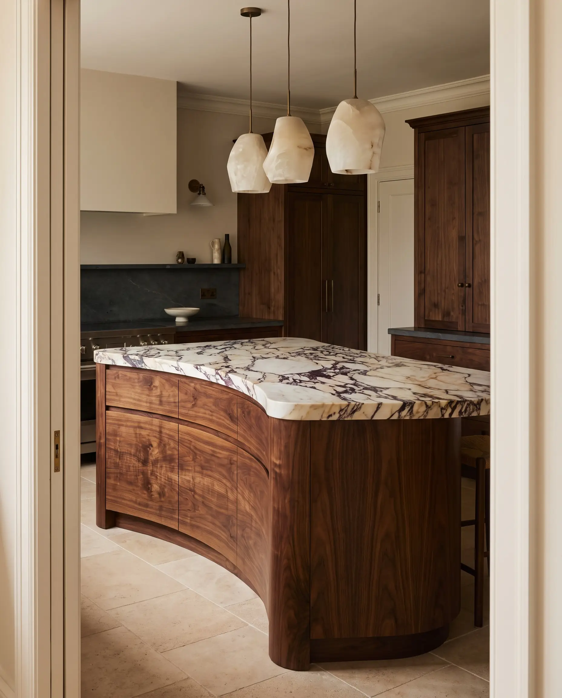 A rounded kitchen island topped with deeply veined Calacatta Viola marble stands over unfilled travertine floors, complemented by warm European walnut joinery and honed soapstone counters.