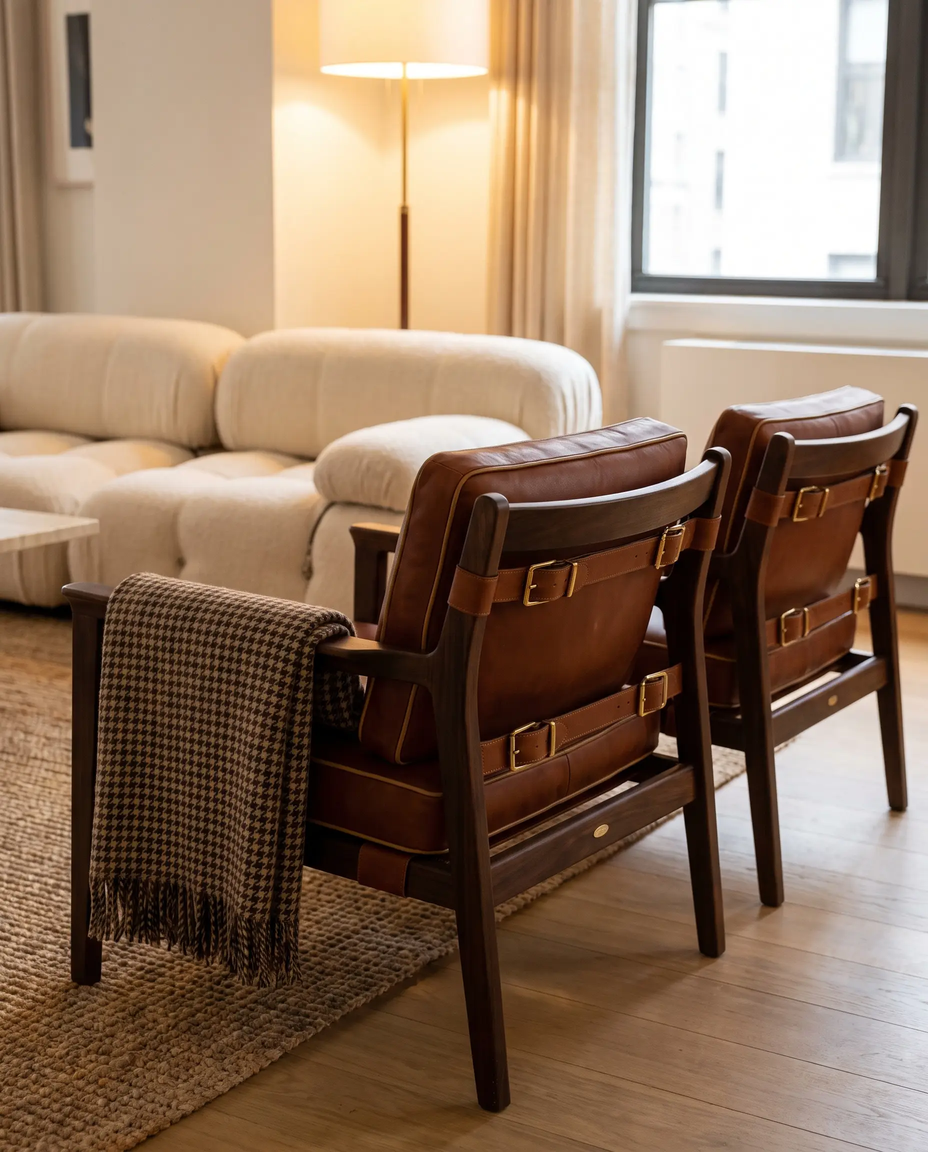 Structured lounge chairs featuring tailored cognac saddle leather and heavy brass buckles sit near an oversized modular sofa and a textured rug.