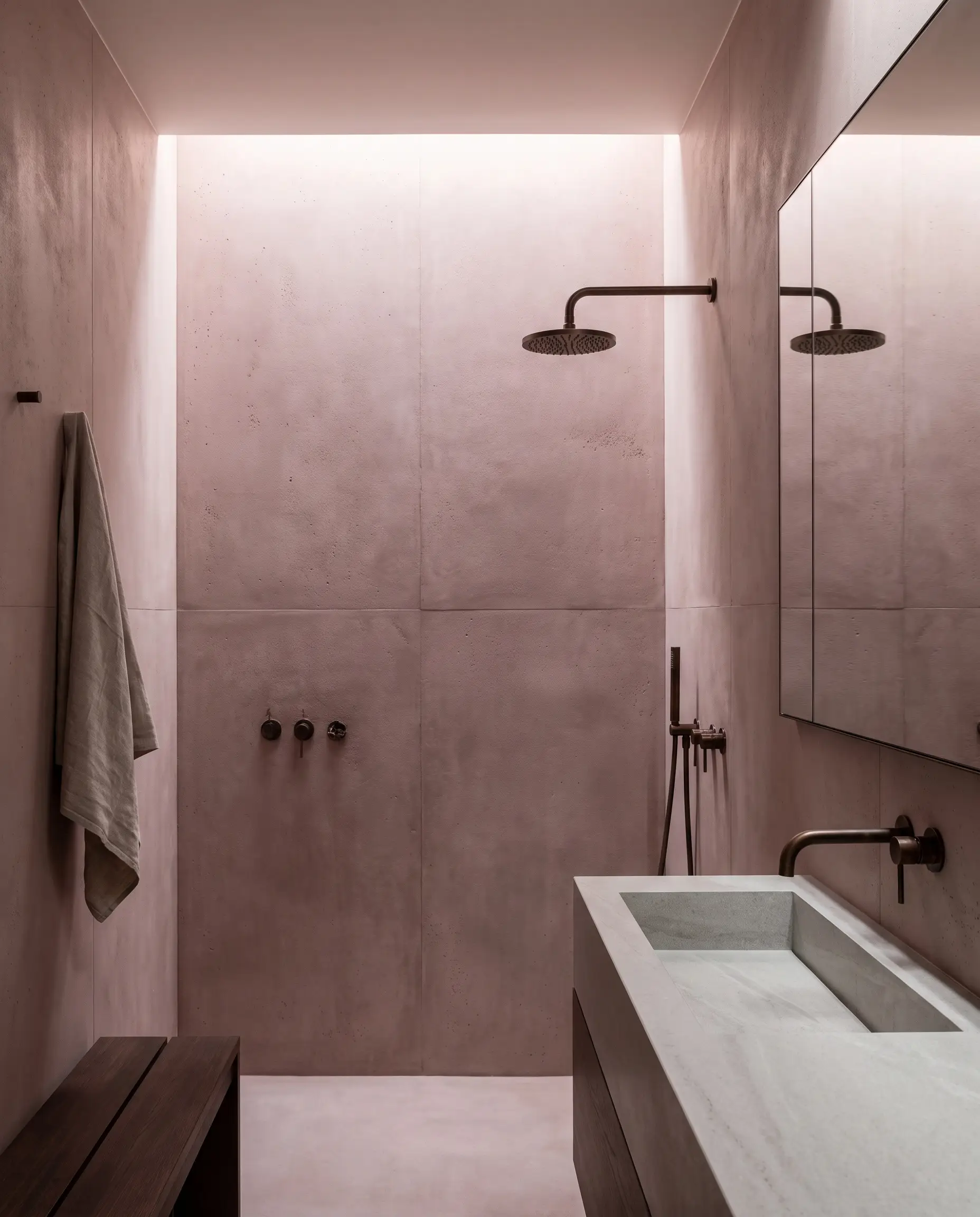 Pigmented dusty rose glass-fiber reinforced concrete panels form a monolithic bathroom wall behind deeply patinated bronze plumbing fixtures and an integrated stone sink.