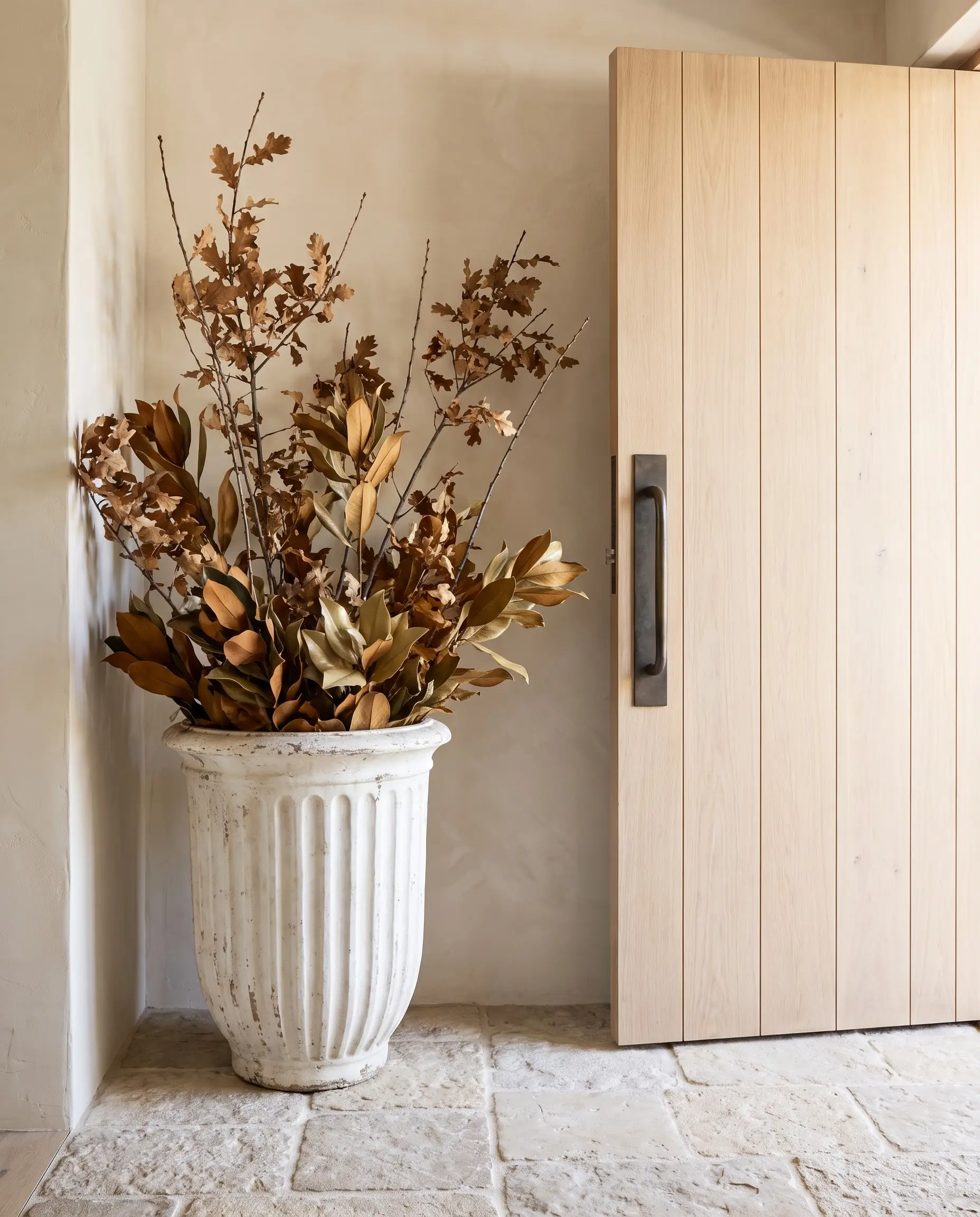 Towering preserved oak branches spring from a heavy fluted Roman plaster planter resting on a tumbled limestone floor beside a white oak door.