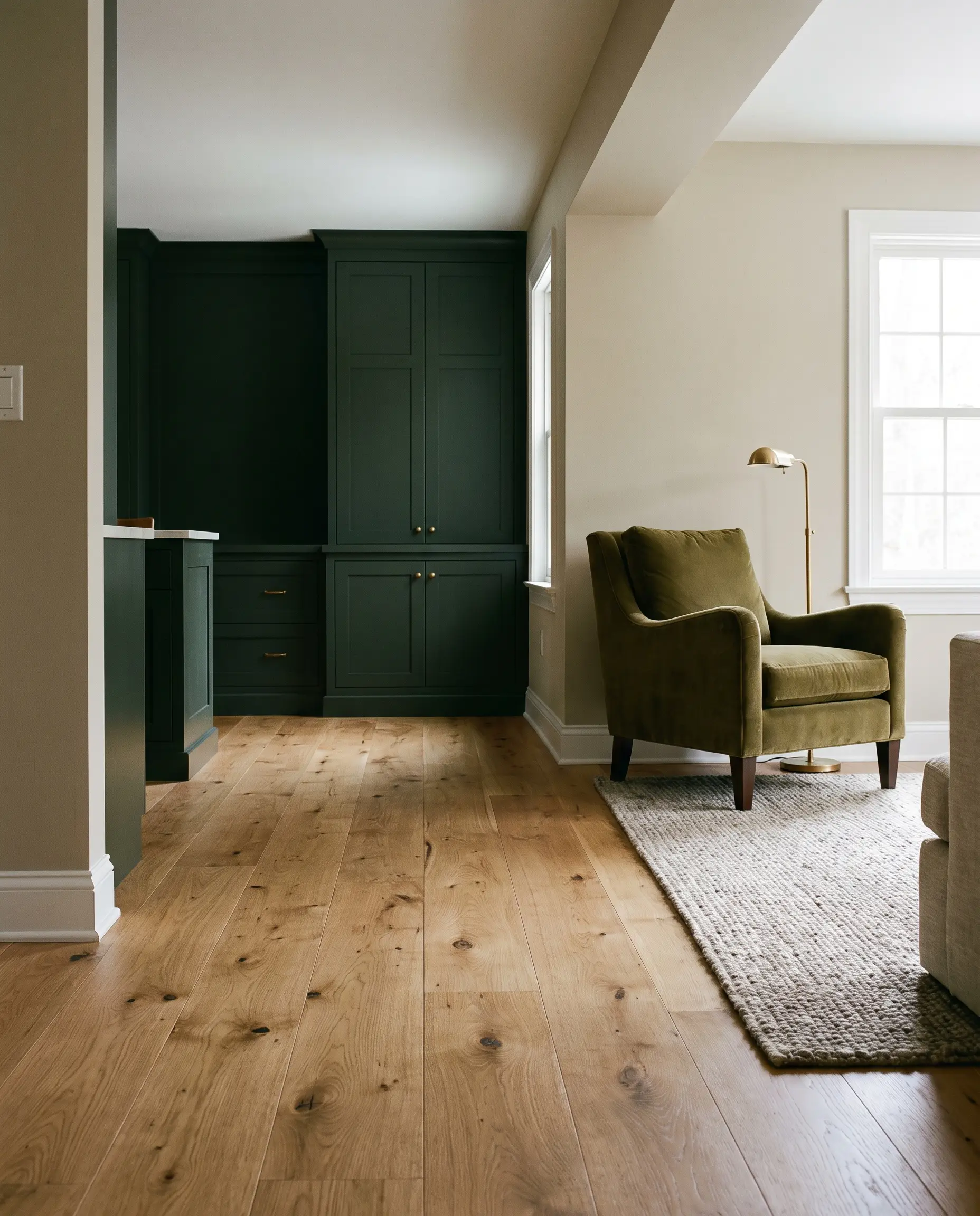 Una poltrona in velluto verde oliva e una lampada in ottone brunito risaltano su listoni in rovere miele, tra pareti nella tonalità Natural Cream di Benjamin Moore e arredi su misura verdi.