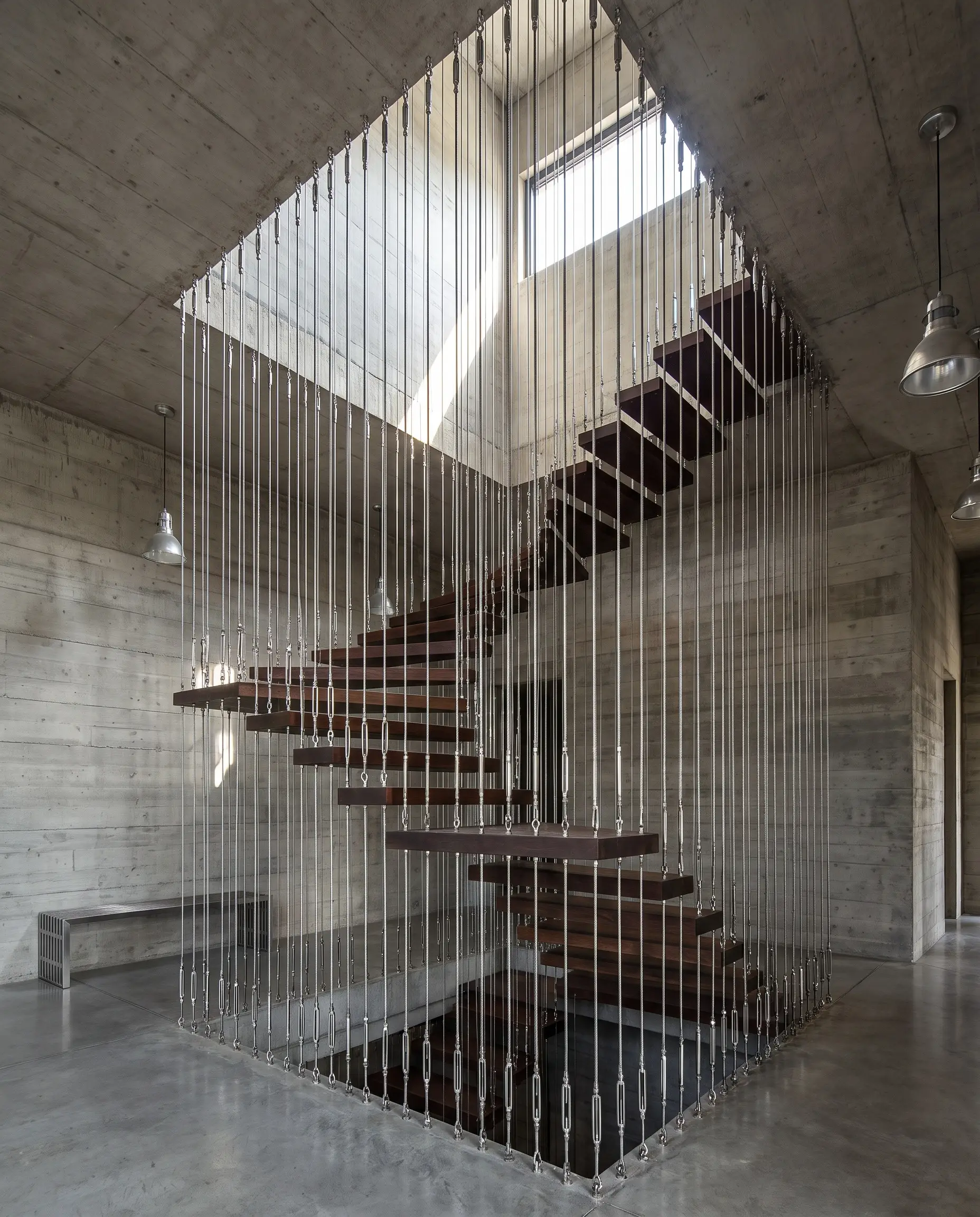 Sleek wooden stair treads are suspended by a geometric cage of floor-to-ceiling marine-grade stainless steel tension cables against exposed concrete walls.