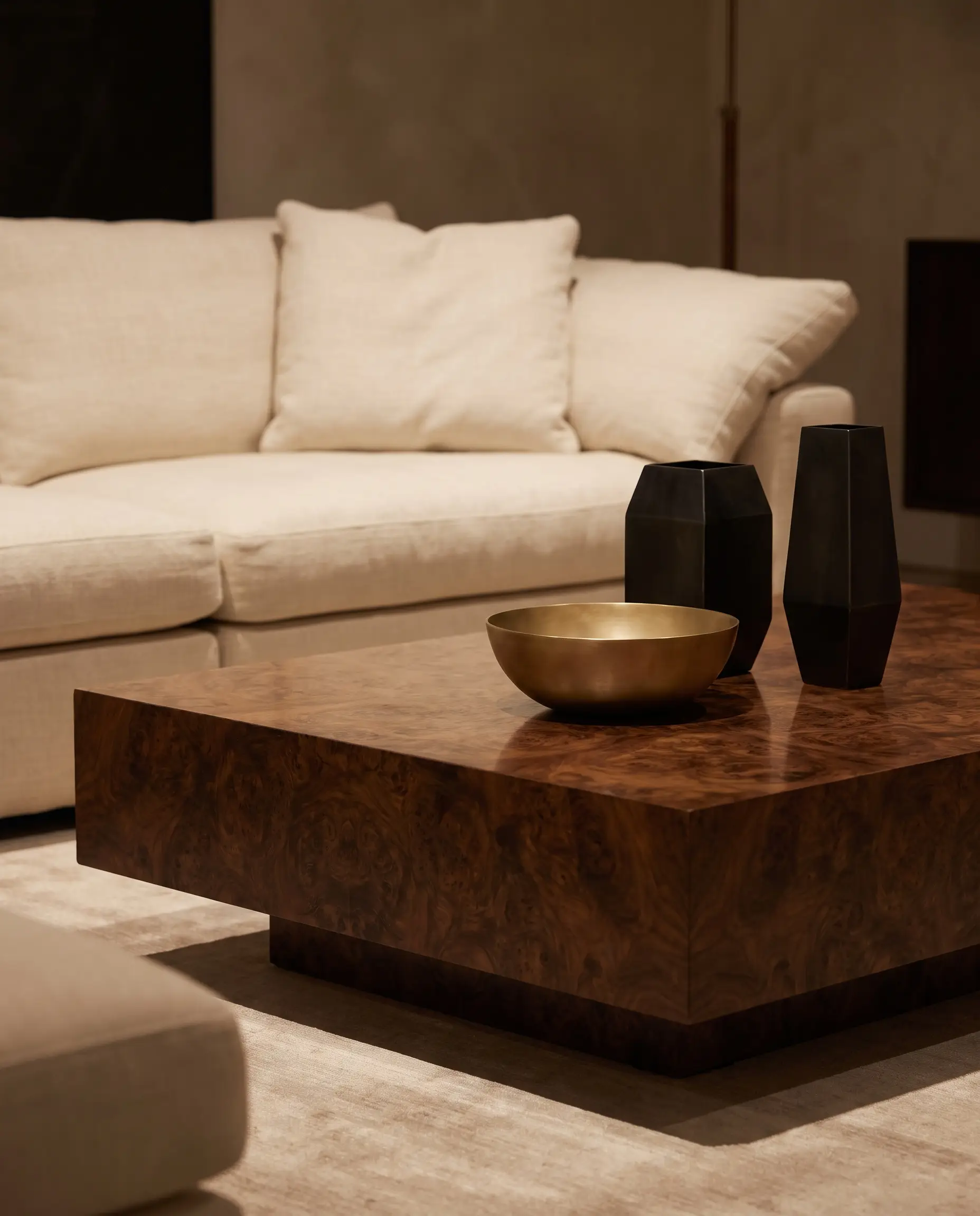 A monumental plinth coffee table crafted from swirling burl wood displays unlacquered brass and blackened steel decor in front of a low-profile cream sofa.