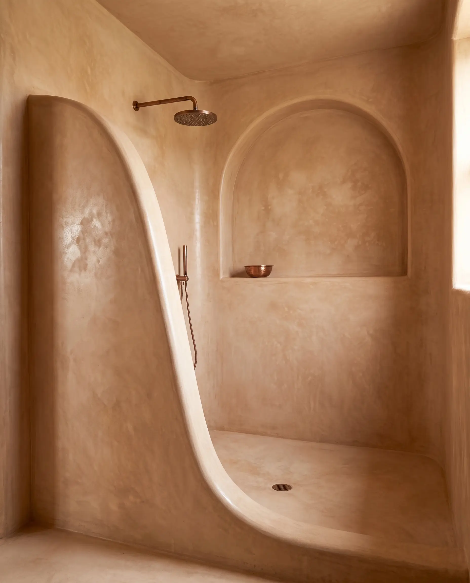 Authentic traditional Moroccan Tadelakt plaster completely covers a sweeping curved shower enclosure and arched architectural niche equipped with minimalist aged copper fixtures.