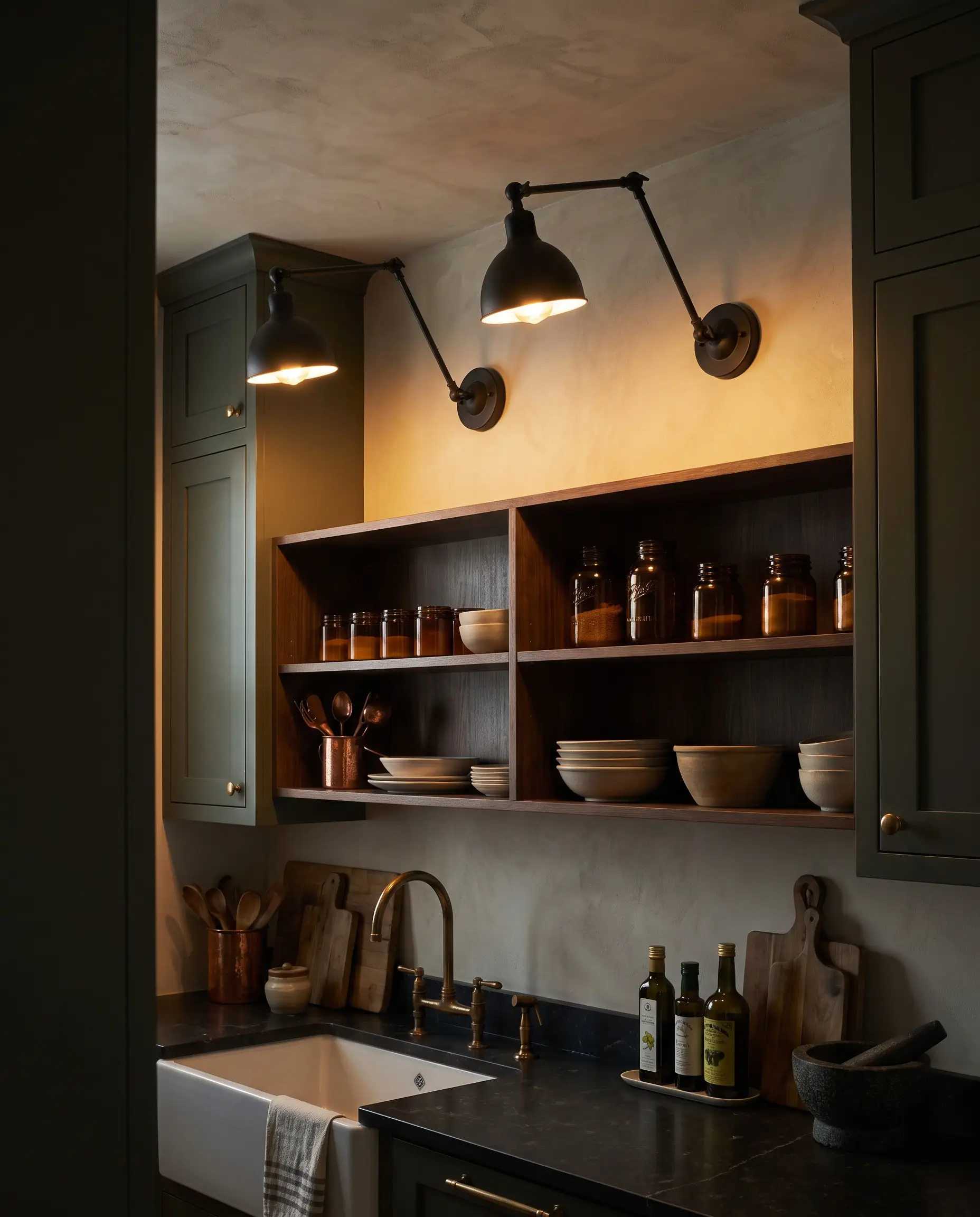 Matte black articulating wall sconces mounted directly above the highest open shelf line illuminate curated jars and culinary tools resting on functional stone prep surfaces.