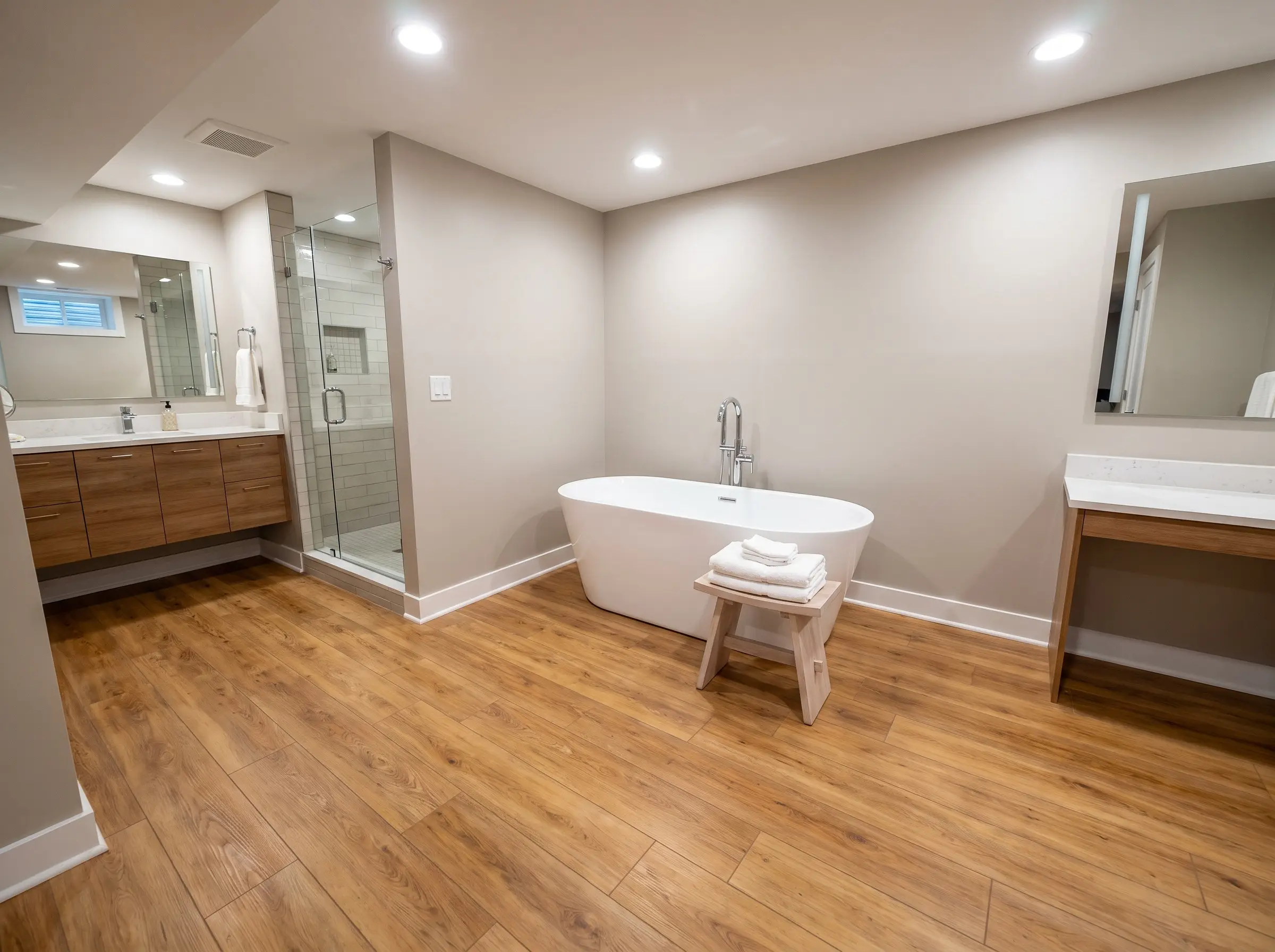 Doghe composite rigide e impermeabili (SPC) portano il calore del parquet in un bagno seminterrato con vasca freestanding bianco candido e sgabello in frassino.