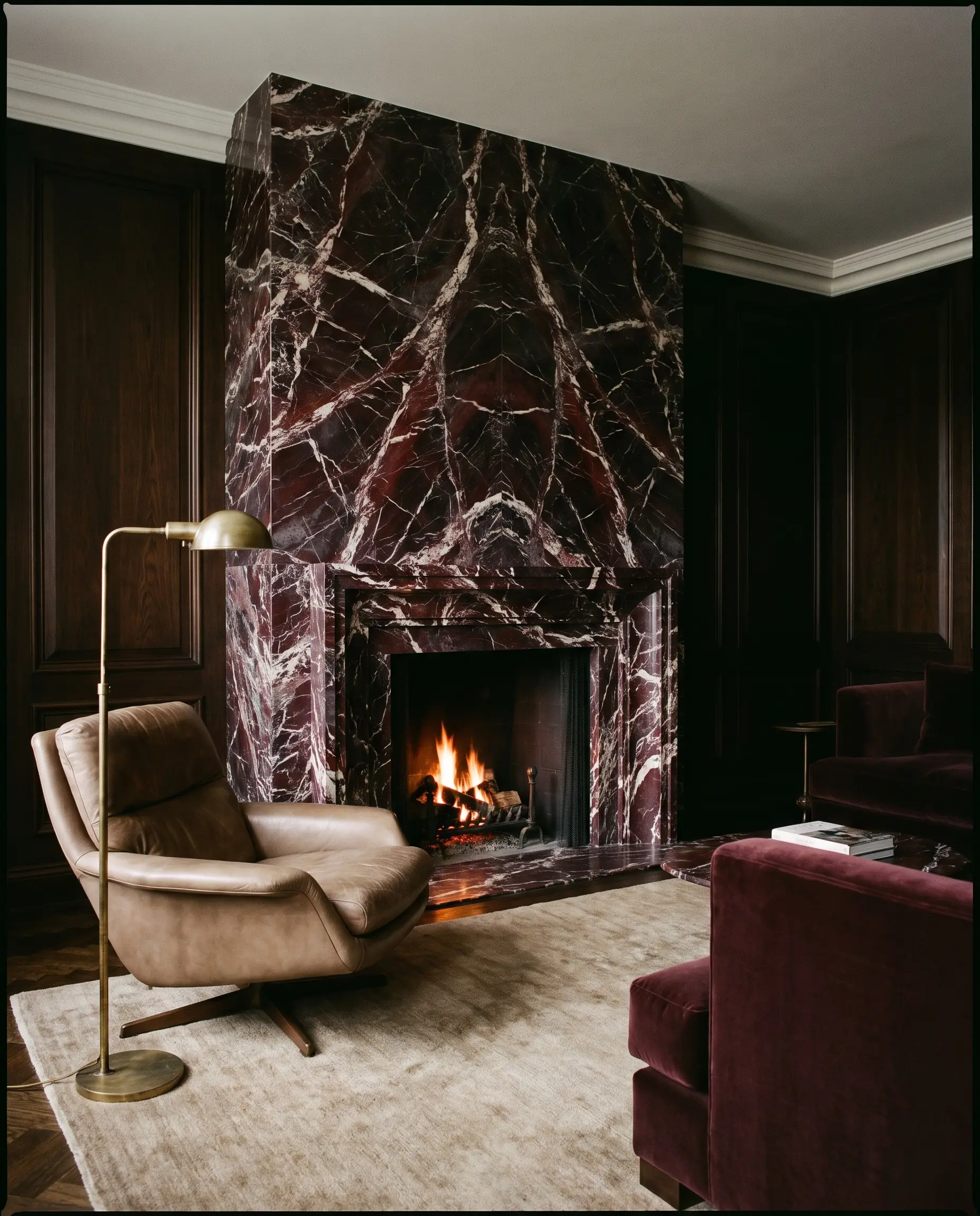 A dramatic fireplace surround constructed from heavily veined Rosso Levanto marble anchors a dark seating area featuring a heavy mushroom leather lounge chair.