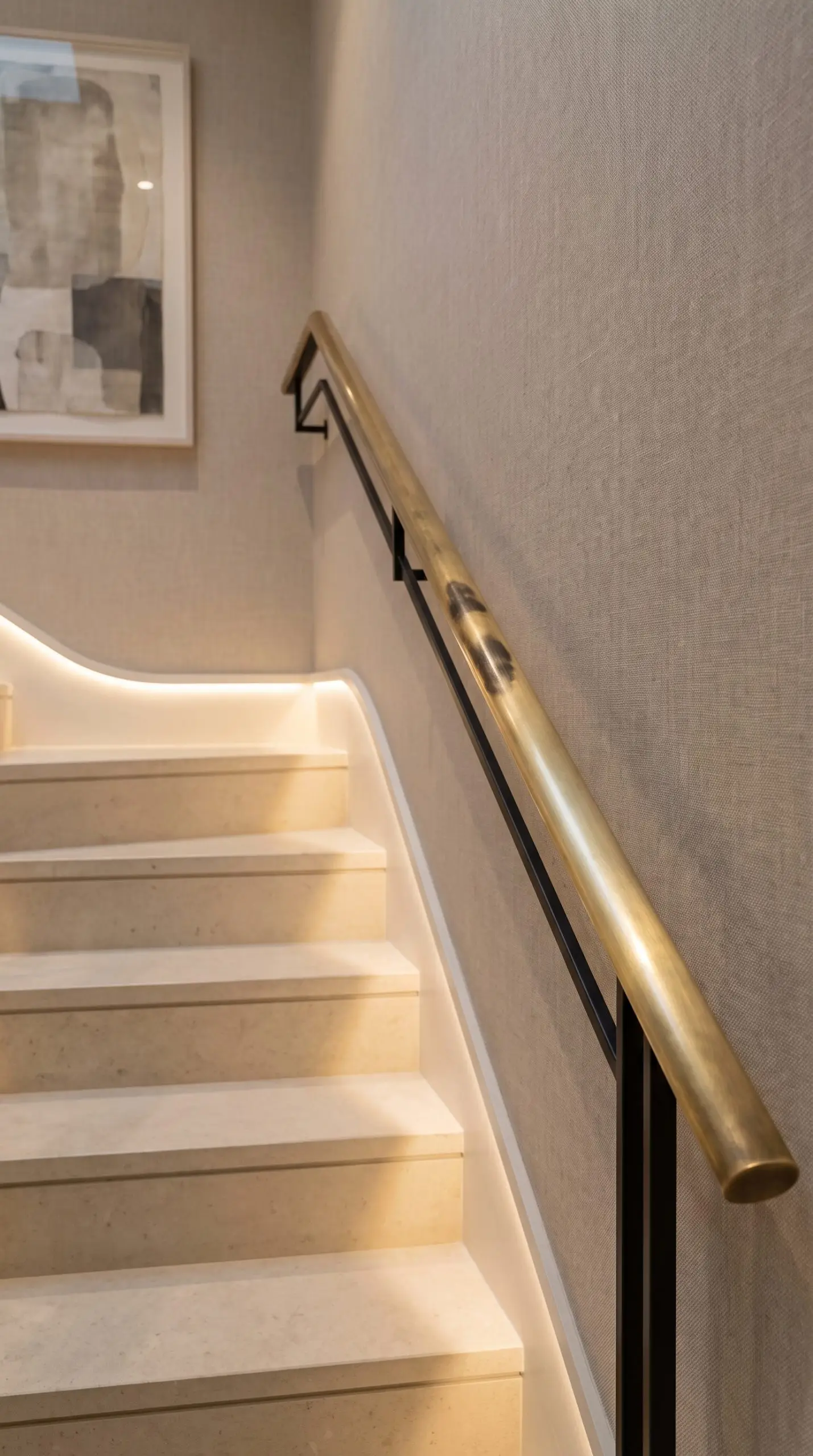 An unlacquered slim-profile brushed brass handrail mounted on minimalist black steel stanchions borders pale stone stair treads against a linen-textured wallcovering.