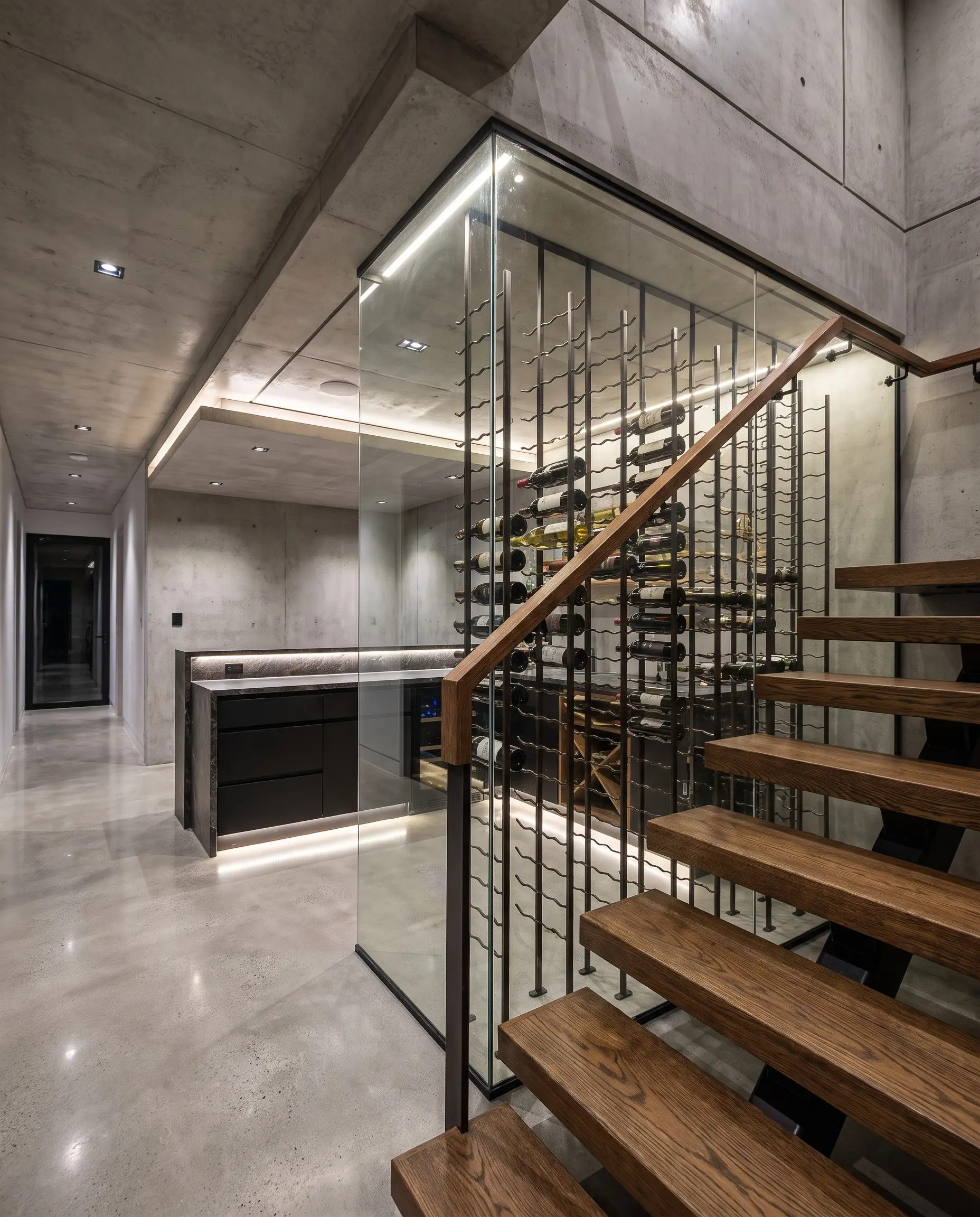 Custom contoured metal racking holds an extensive collection inside a glass-enclosed wine cellar tucked seamlessly beneath heavy structural residential staircase treads.
