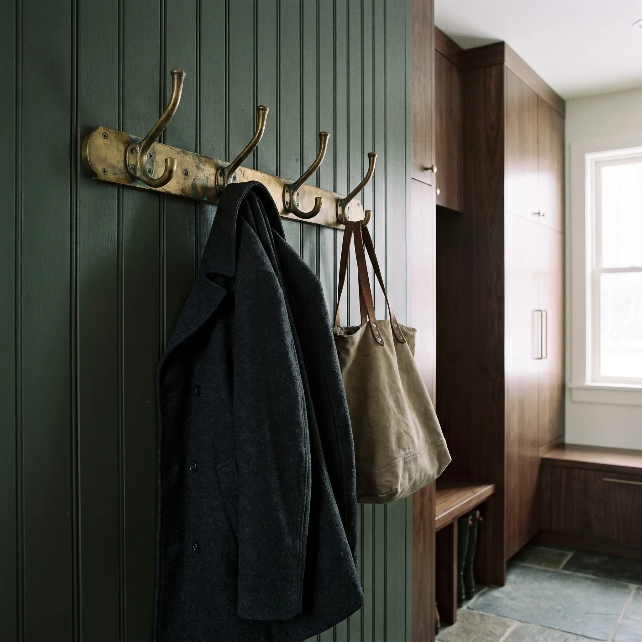 Heavy unlacquered brass utility hooks with a natural bronze patina hold a wool coat against a Sherwin-Williams Rookwood Dark Green wood panel.