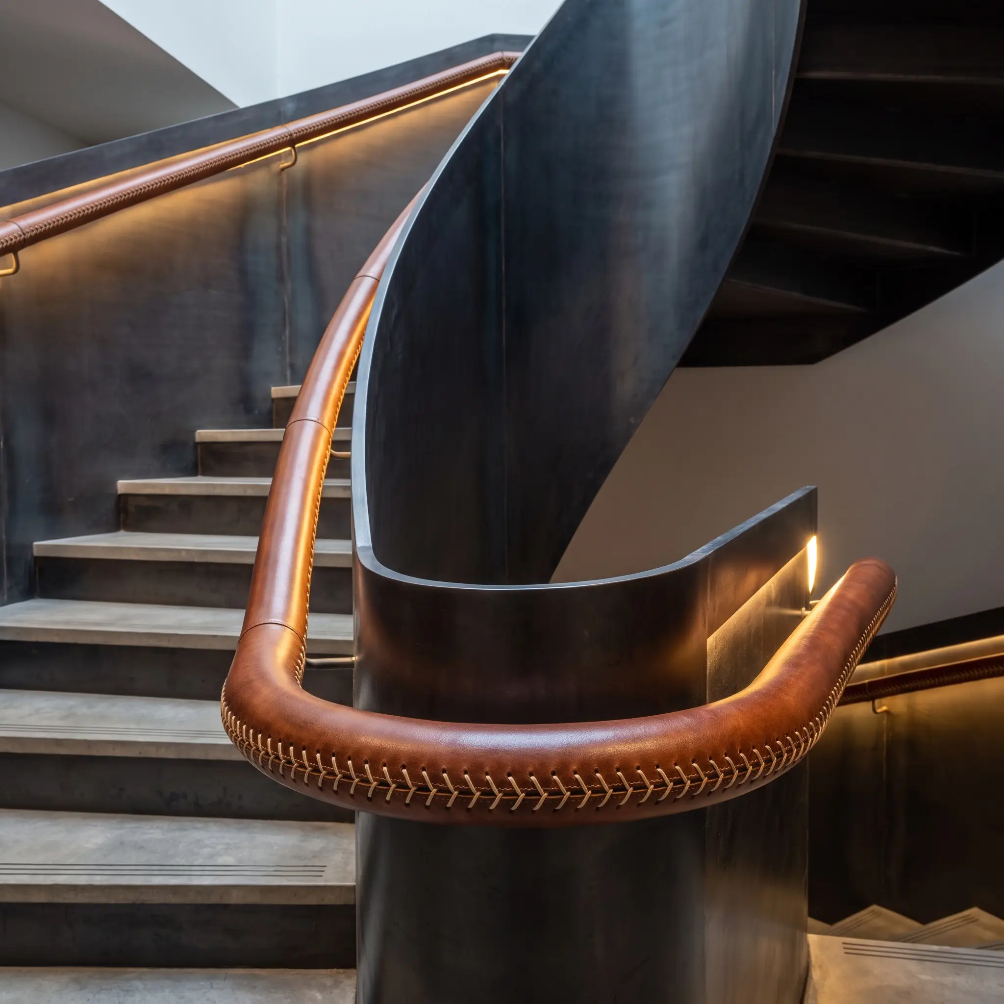 A continuous steel staircase handrail tightly wrapped in premium veg-tanned saddle leather with a precise baseball stitch contrasts against minimalist concrete treads.